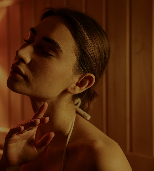 A young woman relaxes inside a warmly lit wooden sauna with her eyes closed and head tilted slightly upward. Soft amber lighting creates a calm, soothing atmosphere, and her serene expression suggests rest and wellness. A bundle of sauna branches or herbs is blurred in the background, adding to the spa-like setting.